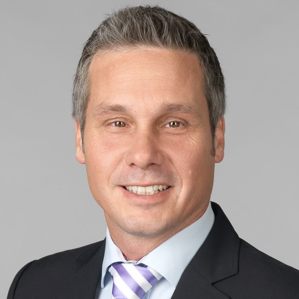 Christian Tschuk filialleiter-in-anzug-mit-krawatte-laechelnd-vor-grauem-hintergrund