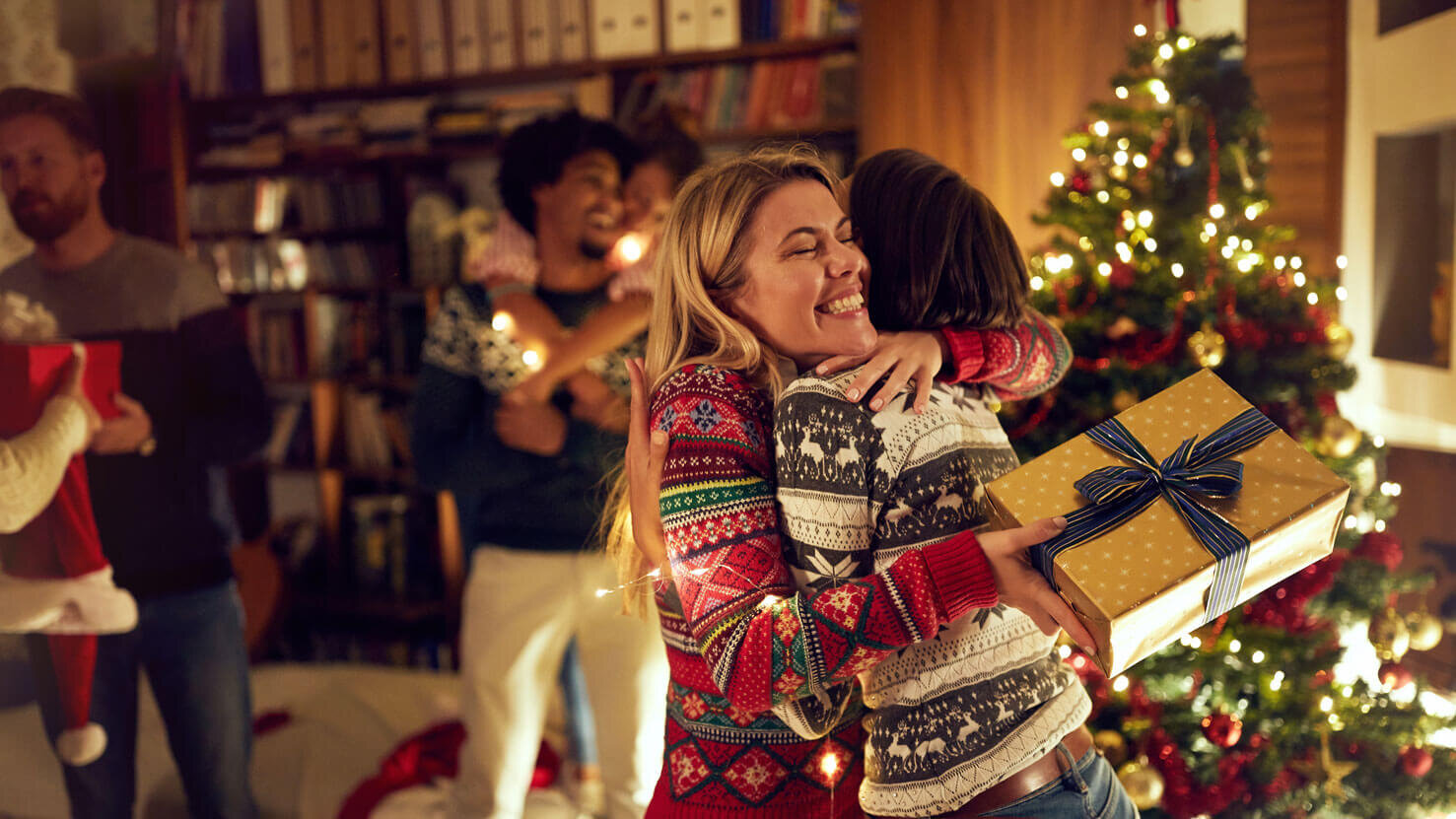 Vintage Schmuck unter dem Weihnachtsbaum- freudige Umarmung zweier Frauen beim Schenken. Im Hintergrund sieht man glückliche Menschen und einen Weihnachtsbaum