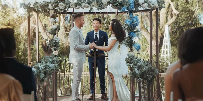 Hochzeitsbudget: Das Bild zeigt eine Frau und einen Mann in Hochzeitskleidung, dieunter einem blumengeschmückten Traubogen stehen und  sich in der Natur das Ja Wort geben