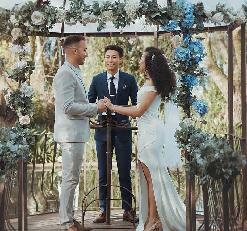Hochzeitsbudget: Das Bild zeigt eine Frau und einen Mann in Hochzeitskleidung, dieunter einem blumengeschmückten Traubogen stehen und  sich in der Natur das Ja Wort geben