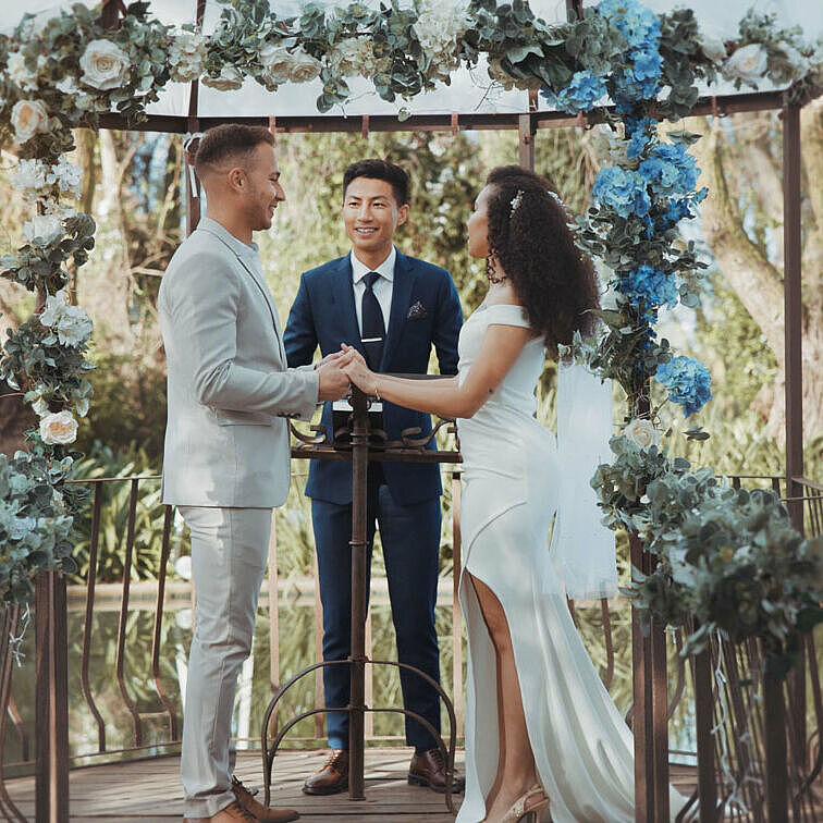 Hochzeitsbudget: Das Bild zeigt eine Frau und einen Mann in Hochzeitskleidung, dieunter einem blumengeschmückten Traubogen stehen und  sich in der Natur das Ja Wort geben
